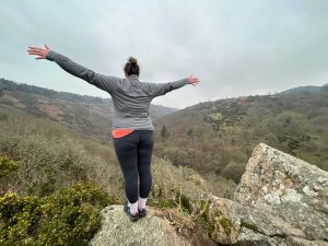 Site owner on top of a Tor on Dartmoor celebrating getting to the top in hiking clothes with arms outstretched.