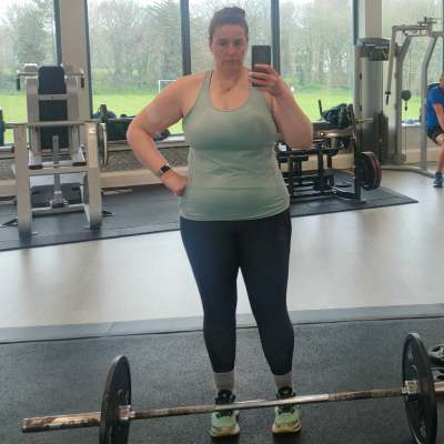 Female in a gym environment with a barbell on the floor in front of her and gym equipment in the background.